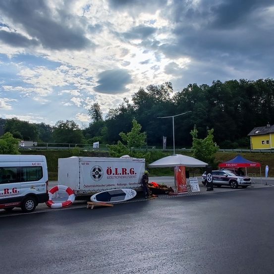 Ein weißer Van und ein weißer Anhänger mit den Buchstaben O.L.R.G. sind am Straßenrand geparkt. Es gibt einen Rettungsring, ein Surfbrett und ein Zelt. Ein Polizeiauto ist in der Nähe geparkt.