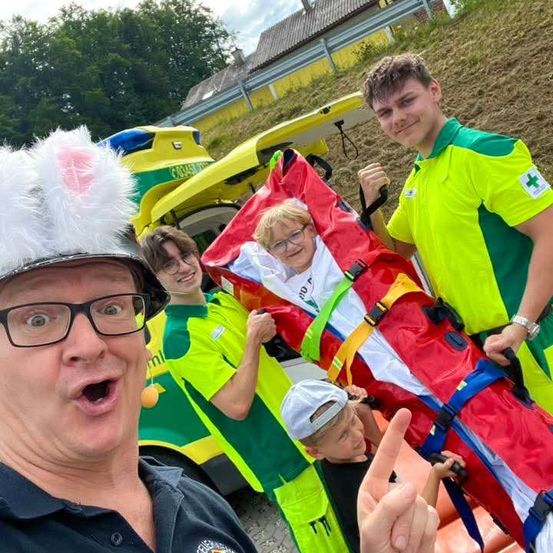 Eine Gruppe von Menschen in grünen und gelben Uniformen trägt eine Trage mit einer Person darin. Die Person vorne trägt einen Hut und eine Brille. Dahinter steht ein gelber Krankenwagen.