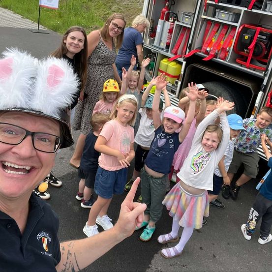 Eine Gruppe von Kindern und Erwachsenen steht vor einem Feuerwehrauto, einige tragen Helme und heben die Hände. Ein Mann in Feuerwehruniform und Mütze mit Hasenohren lächelt und posiert für ein Foto.