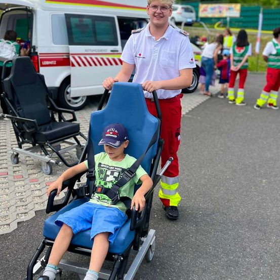 Ein junger Junge im Rollstuhl wird von einem Mann in einem weißen Hemd und roten Hosen geschoben. Sie sind draußen in der Nähe eines Krankenwagens.