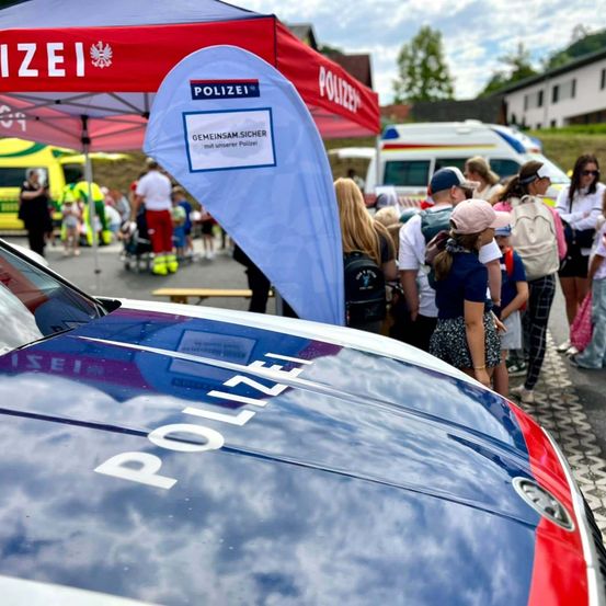 Ein Polizeiauto ist am Straßenrand geparkt, mit einer Menschenmenge, die in der Nähe steht. Im Hintergrund befindet sich ein Polizeizelt, und ein Banner mit dem Wort 'POLIZEI' ist sichtbar.