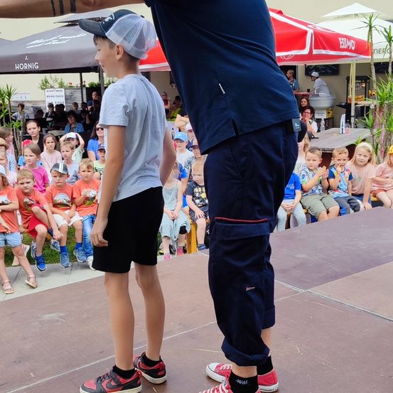 Ein junger Junge steht mit einem Erwachsenen auf der Bühne und beide schauen auf eine Menge von Kindern, die auf Bänken unter Sonnenschirmen sitzen.