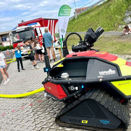 Ein roter und schwarzer Roboter mit dem Wort WOLF an der Seite steht auf dem Bürgersteig. Dahinter beobachten eine Gruppe von Kindern einen Feuerwehrwagen. Eine Fahne mit grünen und weißen Farben befindet sich an der Seite.