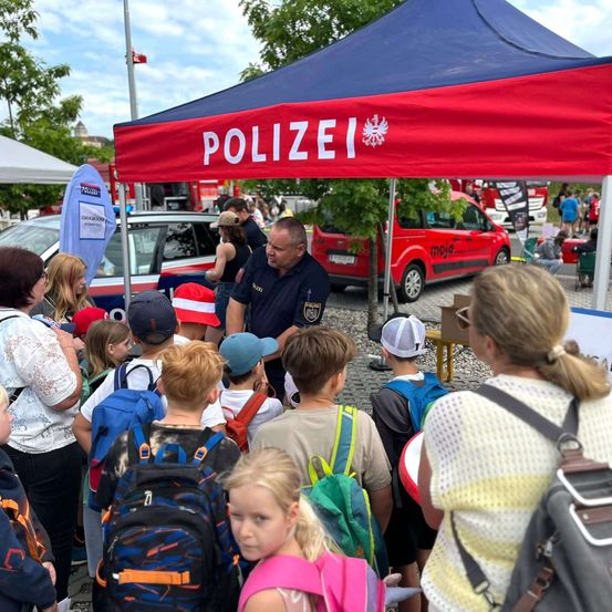 Eine Gruppe von Kindern und Erwachsenen versammelt sich unter einem roten Zelt mit dem Wort Polizei darauf. Ein Polizeibeamter spricht mit den Kindern, und mehrere Rucksäcke sind sichtbar. Im Hintergrund steht ein rotes Rettungsfahrzeug.