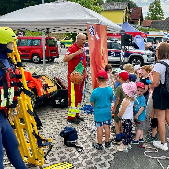 Ein Mann in roter Uniform demonstriert einer Gruppe von Kindern unter einem Zelt eine Rettungstechnik. In der Nähe befindet sich ein Dummy in einem Rettungsweste, eine gelbe Trage und verschiedene Sicherheitsausrüstung.