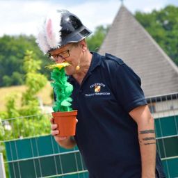 Ein Mann mit Feuerwehrhelm und Hasenohren hält eine Topfpflanze mit gelber Blume und grünen Blättern. Er steht im Freien.
