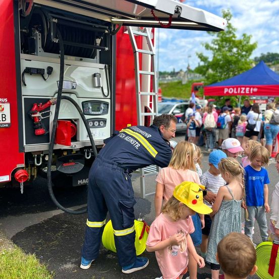 Ein Feuerwehrmann demonstriert einem Kindergruppen das Feuerwehrauto, mit einem roten Feuerlöscher und einer Schlauch auf dem Auto.