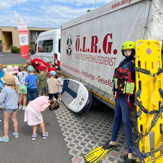 Eine Gruppe von Kindern versammelt sich um einen Krankenwagen, an dem eine Puppe in einem Rettungswesten und Schnorchelausrüstung steht. Der Krankenwagen ist mit dem Text 'O.L.R.G. Katastrophenhilfsdienst' beschriftet.