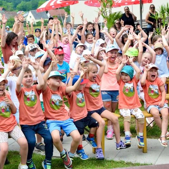 Eine Gruppe von Kindern in passenden orangefarbenen T-Shirts mit grünen Grafiken jubelt und hebt ihre Hände in einem Park.