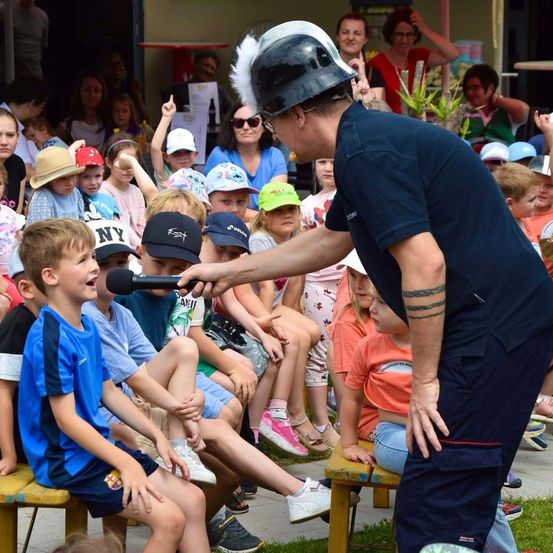 Ein Feuerwehrmann in Uniform hält ein Mikrofon und spricht zu einer Gruppe von Kindern, die auf Bänken sitzen. Die Kinder tragen Mützen und Turnschuhe.
