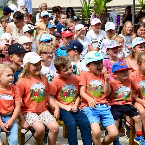 Eine Gruppe von Kindern in passenden orangefarbenen T-Shirts mit Cartoon-Charakteren sitzt auf Bänken, einige tragen Mützen. Sie scheinen bei einer Veranstaltung im Freien zu sein.