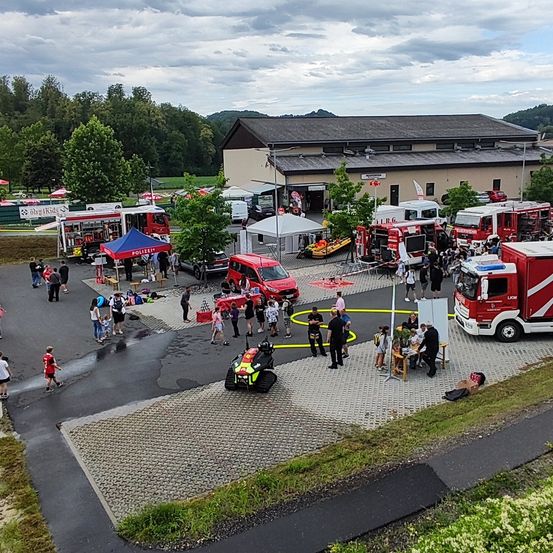Eine Draufsicht auf eine Feuerwehrstation mit mehreren Fahrzeugen und Zelten, bei der sich Menschen versammelt haben, einige gehen und andere stehen in der Nähe der Fahrzeuge.