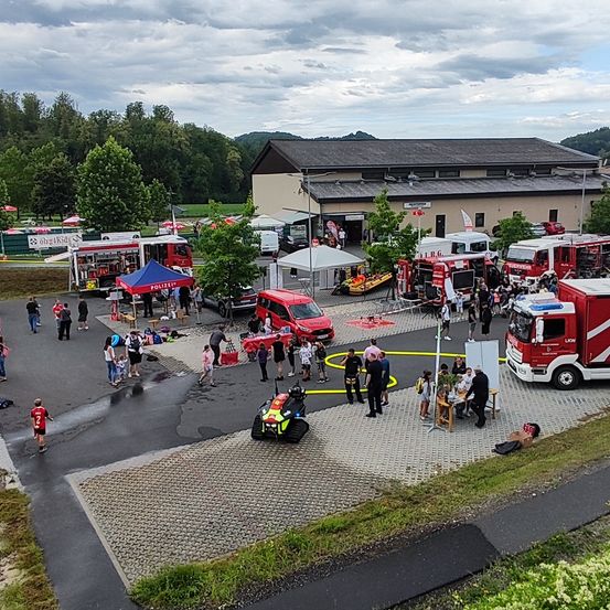 Eine Luftaufnahme einer Feuerwache mit mehreren Feuerwehrautos, die draußen geparkt sind. Menschen gehen herum, einige stehen in der Nähe der Autos und andere versammeln sich um einen Tisch. Es gibt Zelte und ein kleines gelbes Boot in der Nähe.