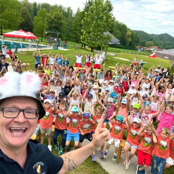 Eine Gruppe von Kindern in passenden orangefarbenen T-Shirts mit Cartoon-Figuren darauf, die in einem Park stehen, während ein Mann mit einem Hasenhut ein Selfie mit ihnen macht.