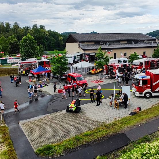 Eine Luftaufnahme einer Feuerwehrstation mit mehreren Feuerwehrwagen und Rettungsfahrzeugen, die draußen geparkt sind. Menschen sind versammelt, einige stehen in der Nähe der Fahrzeuge, während andere herumlaufen. Es gibt Zelte und Tische, die im Bereich aufgestellt sind.