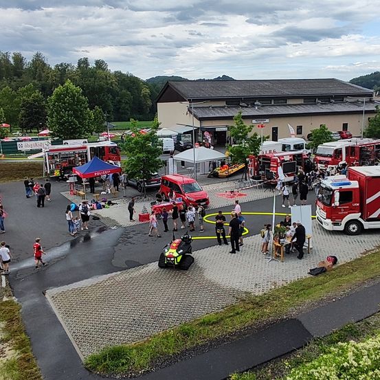 Eine Draufsicht auf eine Feuerwache mit mehreren Feuerwehrautos, die draußen geparkt sind. Mehrere Menschen sind versammelt, einige gehen, und es gibt Zelte und Tische aufgebaut.