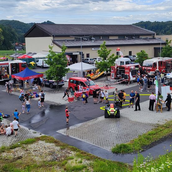 Ein Luftbild einer Gemeinschaftsveranstaltung mit Menschen, die Feuerwehrautos, ein rotes Auto und einen roten Traktor erkunden. Die Veranstaltung findet auf einem Parkplatz mit Zelten, einem Gebäude und einem Berg im Hintergrund statt.