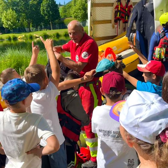 Eine Gruppe von Kindern und ein Mann in einem roten Shirt stehen um ein aufblasbares Boot herum. Der Mann gibt Anweisungen, und die Kinder hören aufmerksam zu.