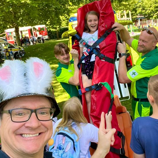 Ein lächelnder Mann in einem Hasenohrenhut macht ein Selfie mit einem Mädchen in einer roten Trage, während zwei Männer in grünen Uniformen ihr helfen. Dahinter sind ein Spielplatz und Feuerwehrwagen zu sehen.