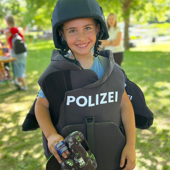 Ein junger Junge, der eine Polizeiweste und einen Helm trägt, lächelt in einem Park, mit Menschen und Bäumen im Hintergrund.