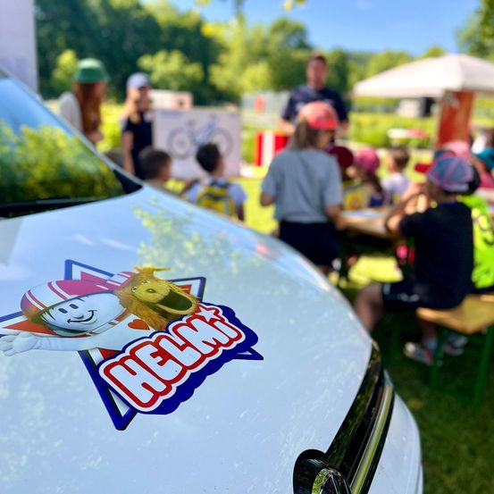 Ein Nahaufnahme des vorderen Stoßfängers eines weißen Autos mit einem bunten Aufkleber, der einen Cartoon-Charakter und das Wort 'Helmt' zeigt. Im Hintergrund sind Menschen um Tische versammelt, die unter einem Schirm stehen.