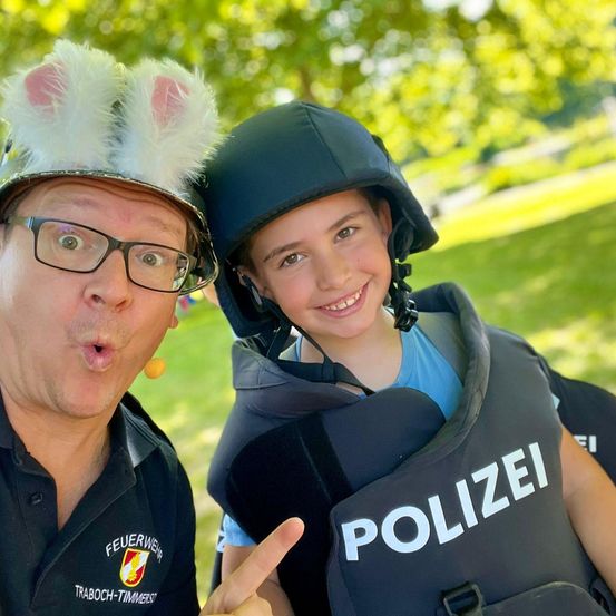 Ein Mann mit einem Hasenohrenhut und Brille posiert neben einem lächelnden Kind in einer Polizeiweste. Sie stehen in einem Park mit Bäumen im Hintergrund.