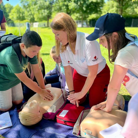 Eine Frau in einem weißen Shirt mit einem roten Kreuz-Patch unterrichtet einen Jungen und ein Mädchen, wie man CPR an einem Mannequin durchführt. Der Junge trägt ein grünes Shirt und das Mädchen eine blaue Mütze. Sie befinden sich auf einem Rasen mit Bäumen im Hintergrund.
