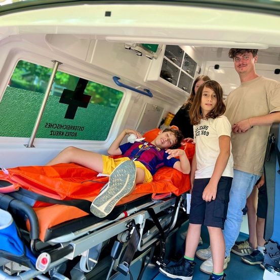 Ein junger Junge liegt auf einer Trage in einem Krankenwagen, während seine Familie in der Nähe steht. Der Junge trägt ein Fußballtrikot, und der Krankenwagen hat medizinische Geräte sichtbar.