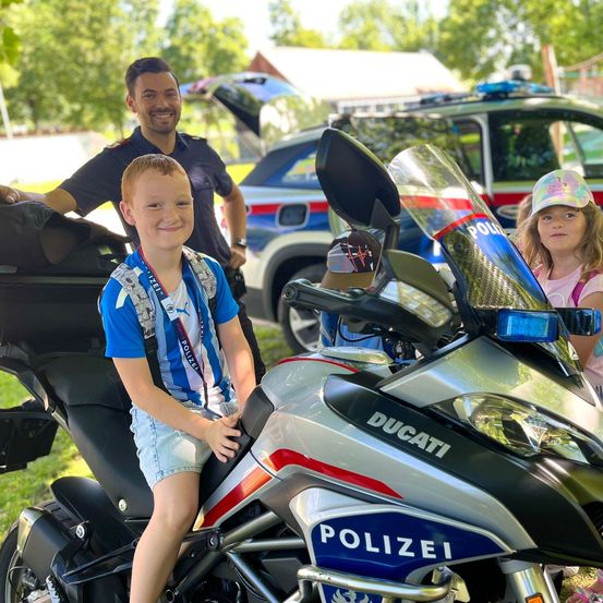 Ein junger Junge sitzt auf einem Polizeimotorrad, lächelt, mit einem Polizisten und einem Mädchen hinter ihm. Das Motorrad ist eine Ducati mit dem Wort 'Polizei' auf der Seite.