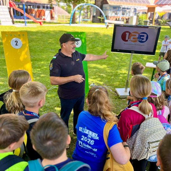 Ein Mann in einem schwarzen Shirt und einer Kappe steht vor einer Gruppe von Kindern und zeigt auf einen Bildschirm, der '1.233' anzeigt. Die Kinder tragen Rucksäcke und scheinen aufmerksam zuzuhören. Der Ort ist im Freien mit einem Spielplatz im Hintergrund.
