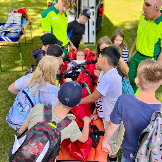 Eine Gruppe von Kindern und Erwachsenen versammelt sich um eine orangefarbene Trage mit einem Baby darauf, möglicherweise in einem Park. Das Baby ist mit Gurten gesichert. Die Kinder tragen Rucksäcke.