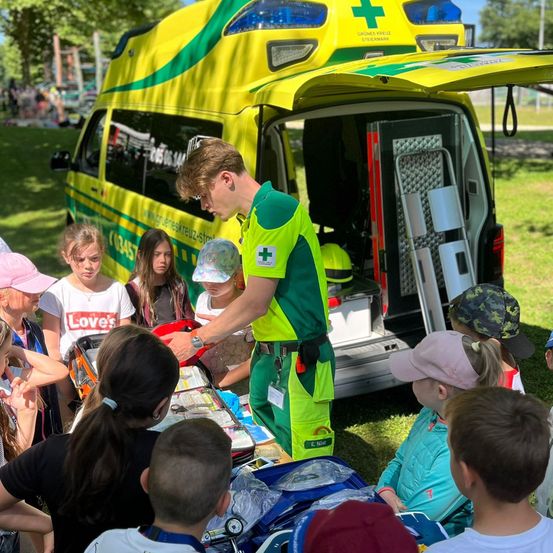 Ein Sanitäter in grüner und gelber Uniform demonstriert Erste-Hilfe-Ausrüstung einer Gruppe von Kindern an einem sonnigen Tag.