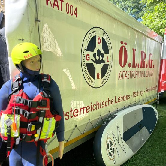 Eine Puppe in einem Neoprenanzug und einer Rettungsweste steht neben einem Rettungsfahrzeug mit der Aufschrift 'O.L.R.G.' und einem Logo. Ein Surfbrett liegt auf dem Rasen in der Nähe.