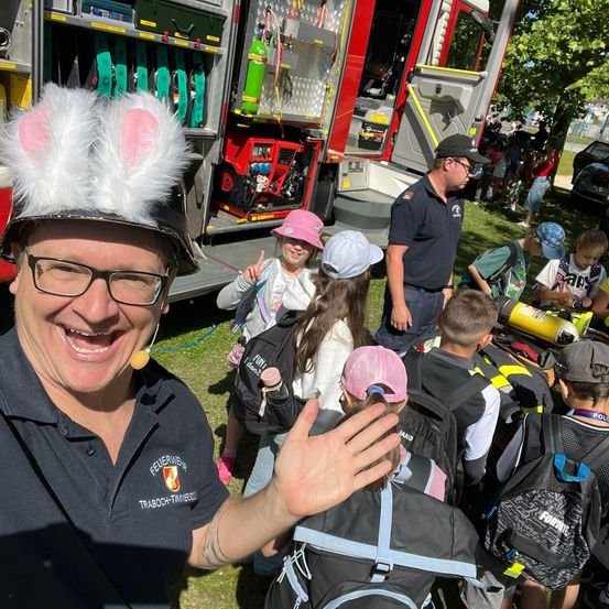 Ein Feuerwehrmann in Hasenohren winkt einer Gruppe von Kindern zu, die vor einem Feuerwehrwagen stehen.