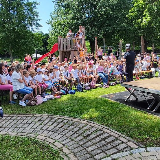 Eine Gruppe von Kindern und Erwachsenen versammelt sich in einem Park, einige sitzen auf Bänken und andere stehen herum. Eine Person mit einem weißen Helm steht in der Nähe eines Tisches. Im Hintergrund befindet sich eine hölzerne Spielstruktur und eine rote Rutsche.