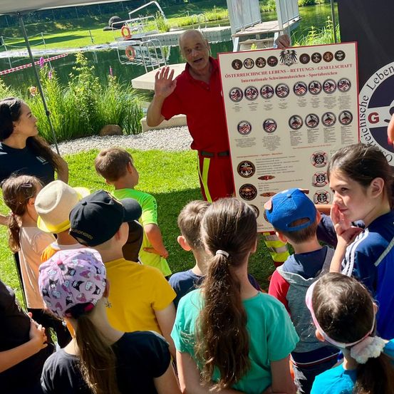 Ein Mann in einem roten Outfit hält ein Poster mit verschiedenen Abzeichen und Symbolen und erklärt etwas einer Gruppe von Kindern in einem Grasbereich in der Nähe eines Sees.