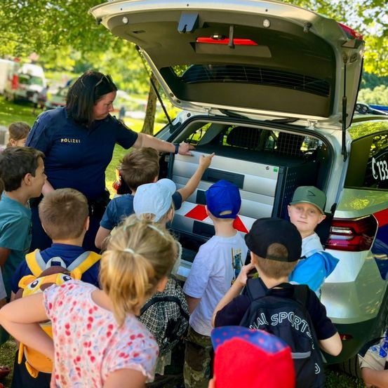 Eine Polizistin zeigt einer Gruppe von Kindern den Kofferraum eines Polizeiwagens, wobei der offene Kofferraum Ausrüstung und einen Käfig offenbart.