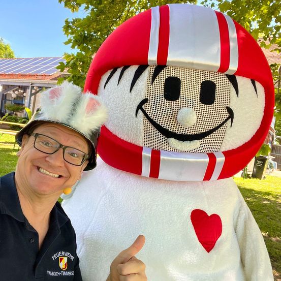 Ein Mann in einem schwarzen Shirt mit 'Feuerwehr' steht neben einem großen Maskottchen, das einen roten und weißen Helm mit einem Herz auf der Brust trägt.