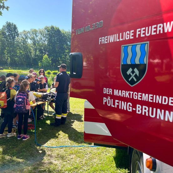 Eine Gruppe von Kindern und Erwachsenen versammelt sich um einen Feuerwehrwagen mit dem Schild 'Freiwillige Feuerwehr'. Der Feuerwehrmann erklärt der Gruppe etwas.