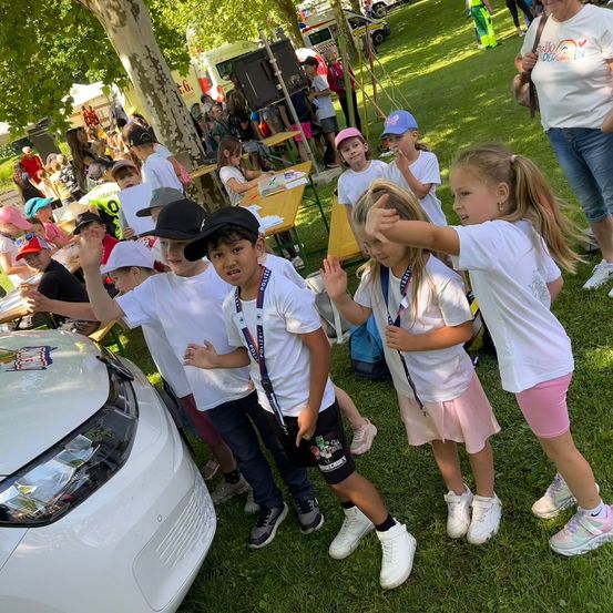 Eine Gruppe von Kindern in weißen Shirts und Sneakers steht um ein weißes Auto in einem Park, mit einer Frau im Hintergrund.