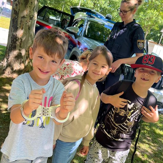 Drei Kinder stehen in einem Park mit einer Polizistin, eines hält Handschellen. Hinter ihnen ist ein Polizeiauto geparkt, in dem eine Frau sitzt.