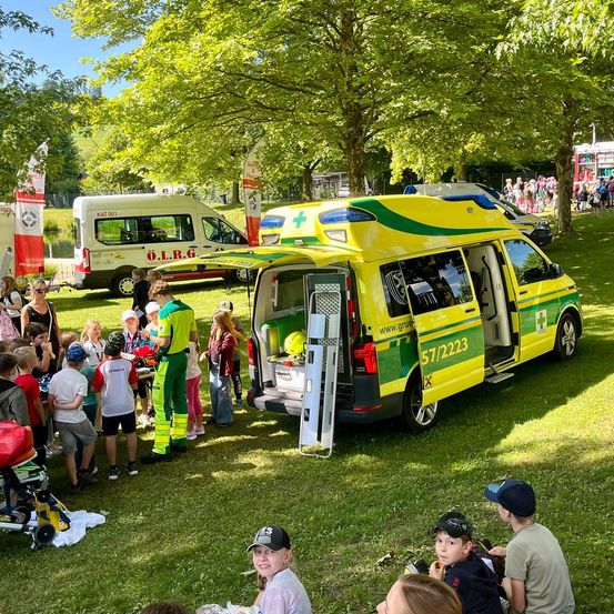 Eine Gruppe von Kindern und Erwachsenen versammelt sich um einen offenen Krankenwagen auf einem Rasenfeld mit Bäumen im Hintergrund.