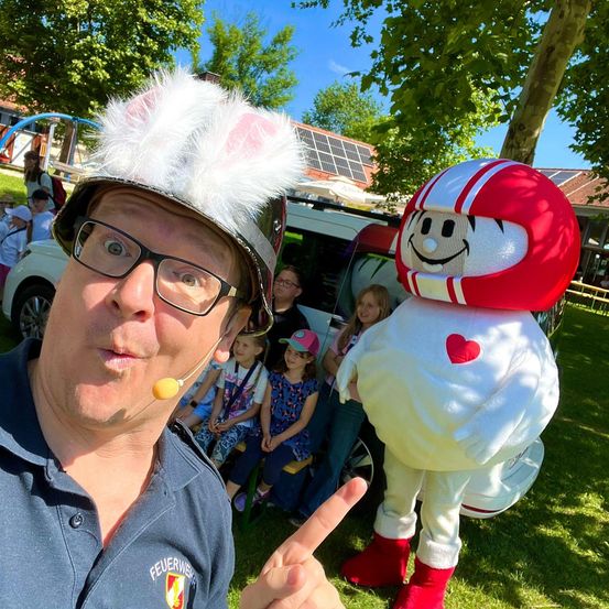 Ein Mann in einem Feuerwehrhelm und Brille posiert mit einer großen Maskottchen-Figur in einem roten Helm und weißem Outfit mit einem roten Herzen darauf, mit Kindern und einem Auto im Hintergrund.