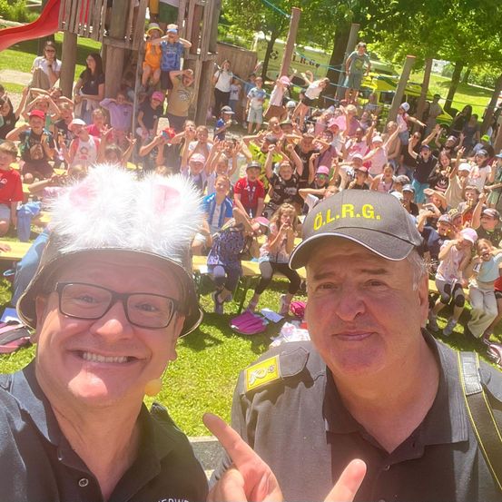 Zwei Männer in Uniform posieren für ein Foto mit einer Menschenmenge von Kindern in einem Park. Ein Mann trägt einen Helm mit Hasenohren.