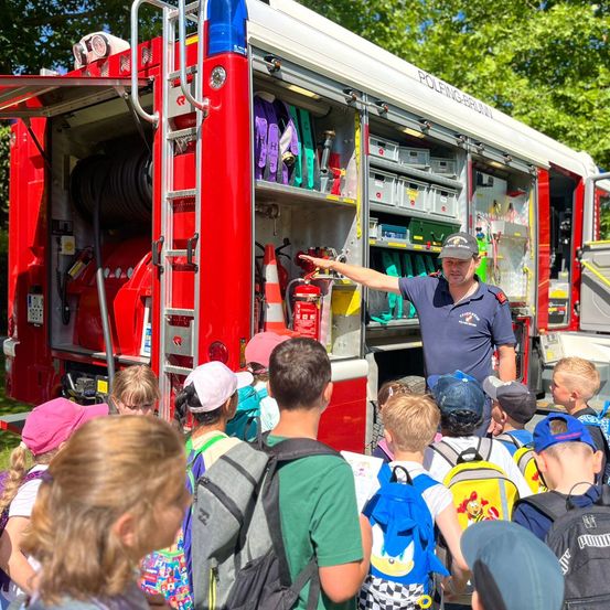 Ein Feuerwehrmann zeigt einer Gruppe von Kindern das Innere eines Feuerwehrwagens, wobei verschiedene Ausrüstungsgegenstände sichtbar sind.