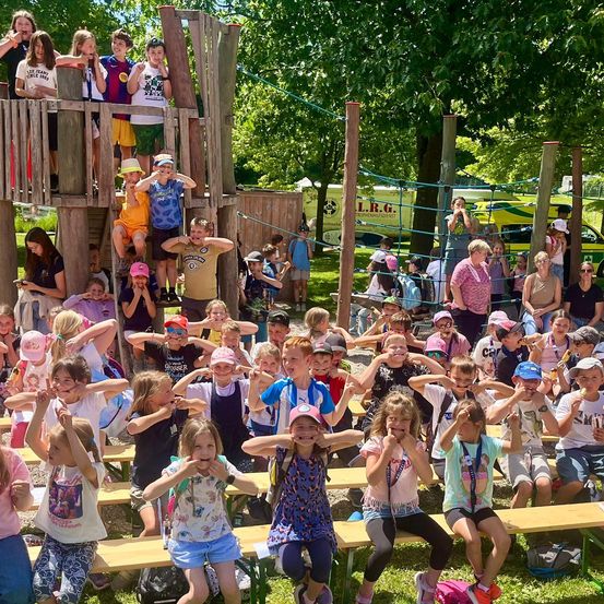 Eine Gruppe von Kindern und Erwachsenen versammelt sich im Freien, einige sitzen auf Bänken, während andere stehen und auf einer Holzspielstruktur klettern. Die Kinder tragen Hüte und einige halten Eis. Im Hintergrund steht ein gelbes Fahrzeug und ein Baum.