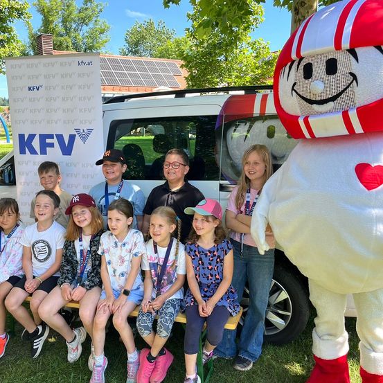 Eine Gruppe von Kindern und zwei Erwachsenen sitzen auf einer Bank vor einem weißen Van mit Solarmodulen und einem Maskottchen. Die Kinder tragen Umhänge und einige haben Baseball-Kappen.