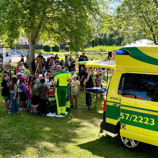 Eine Gruppe von Kindern und Erwachsenen versammelt sich um einen Krankenwagen mit offener Hecktür, möglicherweise für eine Demonstration oder Veranstaltung.