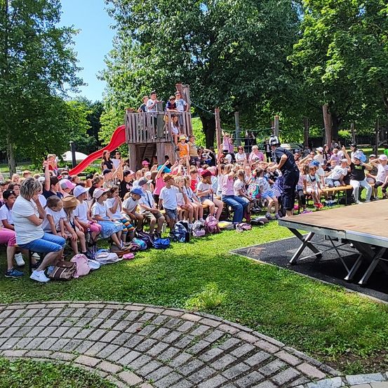 Eine Gruppe von Kindern und Erwachsenen versammelt sich in einem Park. Einige sitzen auf dem Gras, während andere stehen und einen Mann auf der Bühne beobachten. Im Hintergrund befindet sich eine Rutsche und eine Bank.