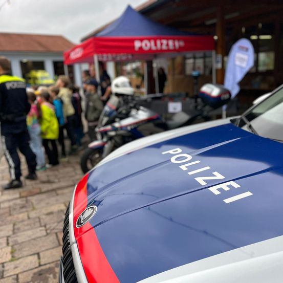 Ein Polizeiauto mit dem Wort 'POLIZEI' auf der Motorhaube steht vor einem Gebäude mit einem Zelt. Mehrere Menschen, darunter Kinder, stehen herum, und ein Motorrad ist in der Nähe geparkt.